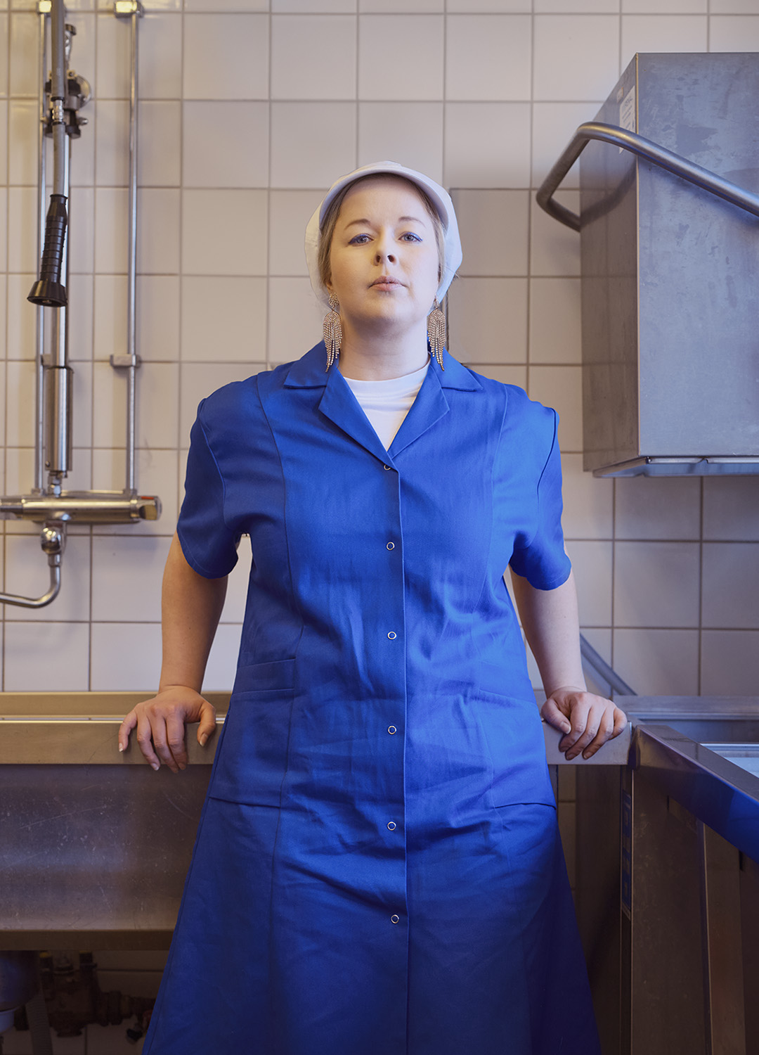 Kvinna i vit keps strassörhängen och blå arbetsrock står lutat mot en diskbänk. Bakom vitt kakel, industridiskmaskin och en vattenkran. 