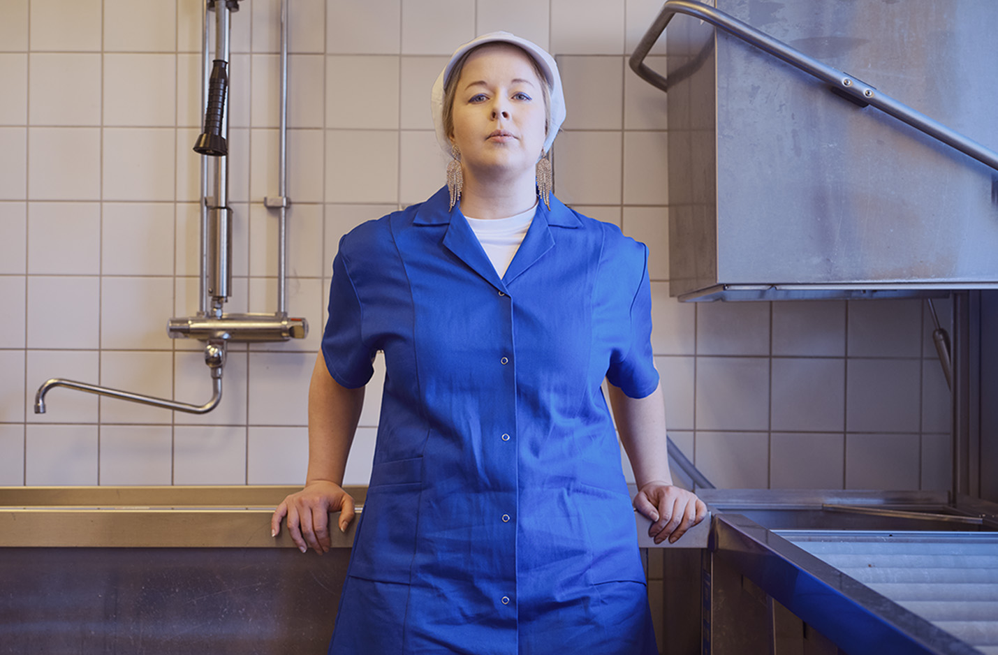 Klickbar bild Kristinvariationer. Kvinna i vit keps strassörhängen och blå arbetsrock står lutat mot en diskbänk. Bakom vitt kakel, industridiskmaskin och en vattenkran. 