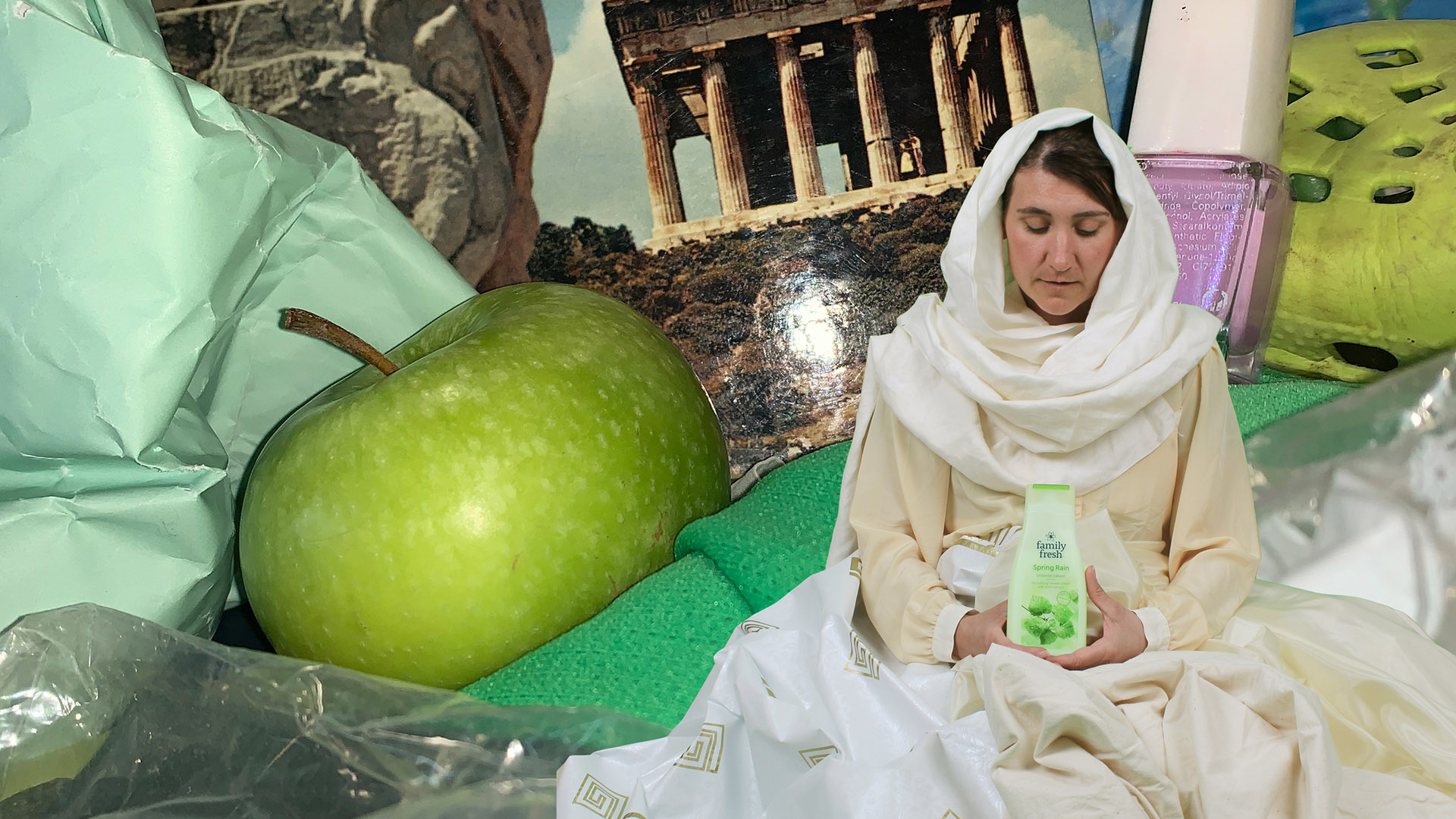 Fotokollage över olika föremål i ovanliga proportioner till varandra. En kvinna i ljus toga och sjal runt huvudet i förgrunden håller i en grön flaska med duschtvål. I barkunden ett enormt grönt äpple, ett hopskrynklat mintgrönt papper, ett vykort föreställande Akropolis, en gigantisk flaska med rosa nagellack och en starkt grön foppatoffla.  