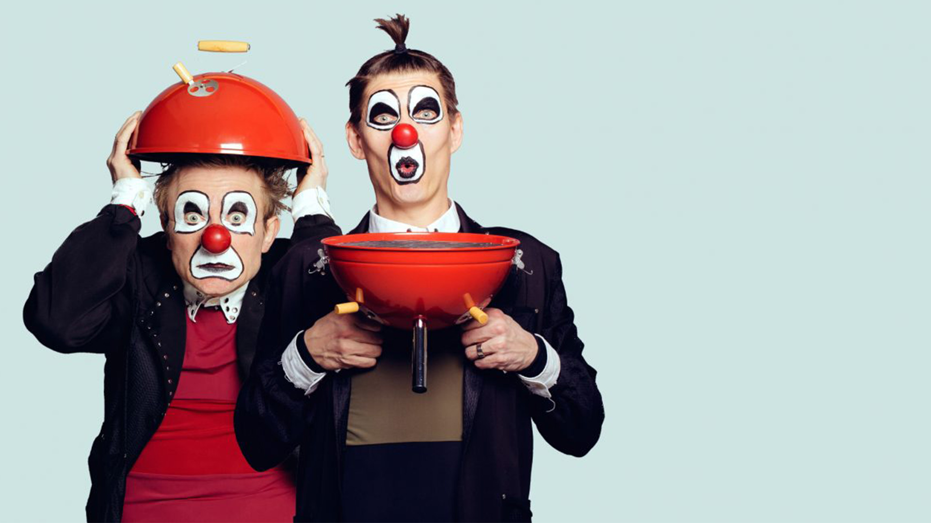 Två clowner i svarta kavajer står framför en turkos bakgrund. De håller en liten röd klotgrill i händerna. Den ena håller locket över huvudet. Den andra underdelen av grillen framför sig nedanför hakan. 