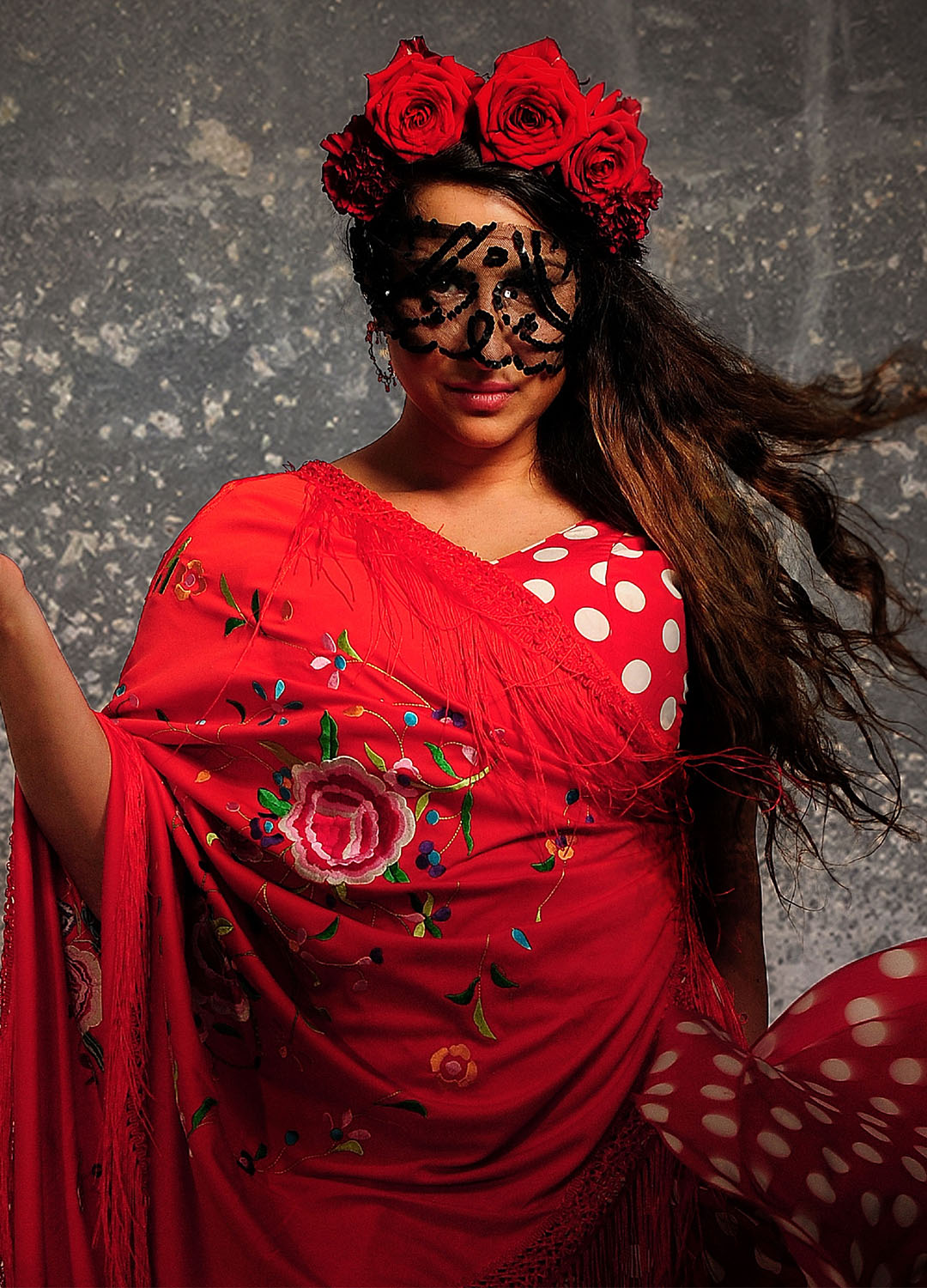 Kvinna i röd klänning med röda blommor i håret och svart spets framför ögonen. Hon håller handen i en danslikande rörelse.