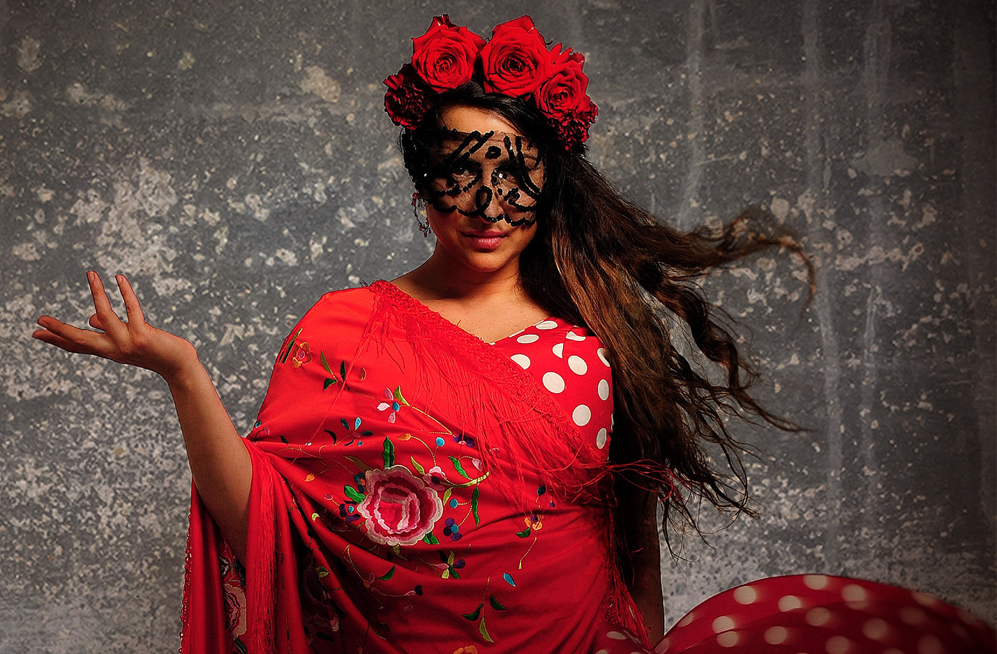 Klickbar bild Carmen med förhinder. Kvinna i röd klänning med röda blommor i håret och svart spets framför ögonen. Hon håller handen i en danslikande rörelse.