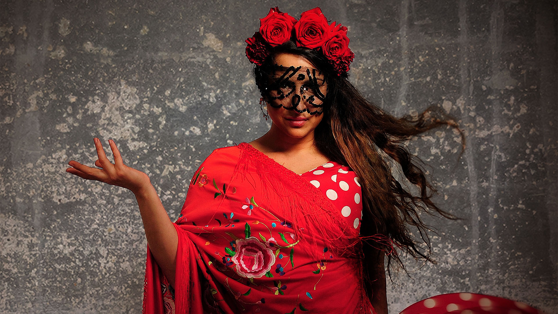 Kvinna i röd klänning med röda blommor i håret och svart spets framför ögonen. Hon håller handen i en danslik rörelse.